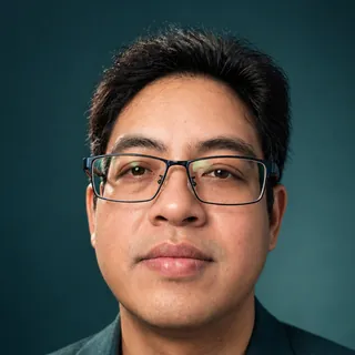 Suparerg Suksai, photographed head-on against a dark teal background, wearing rectangular glasses and a dark jacket.
